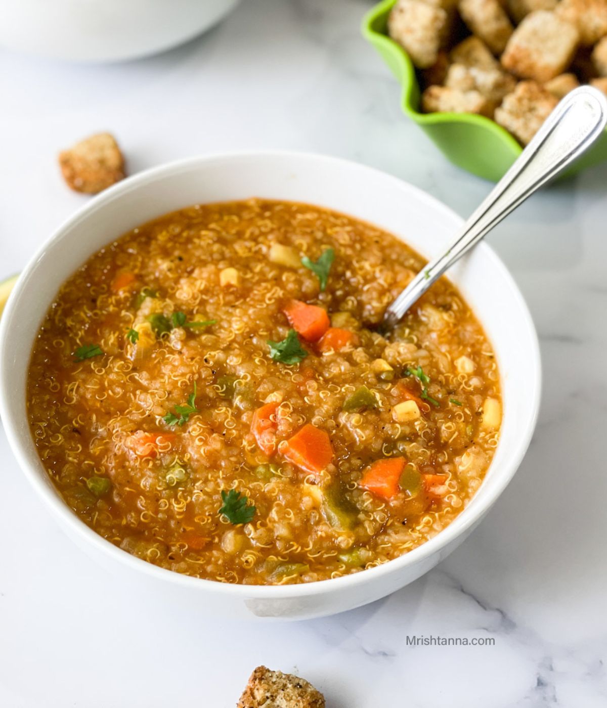 A white bowl is with Quinoa vegetable soup and spoon is inserted.