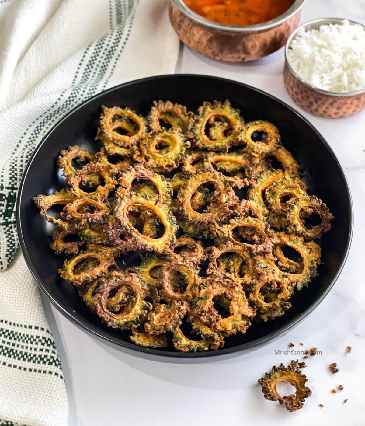 A large plate is with air fryer karela chips and bowl of rasam and rice on the side.