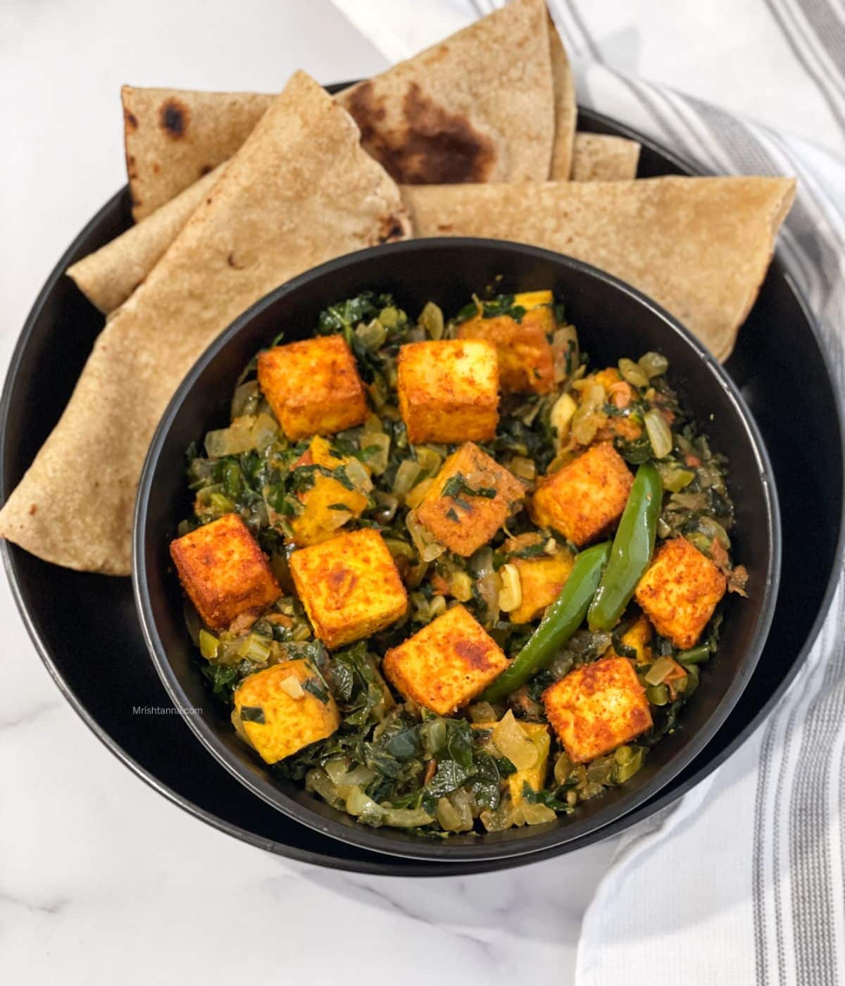 A plate is with chapati and bowl of tofu saag paneer on the table with cloth napkins by the side. 
