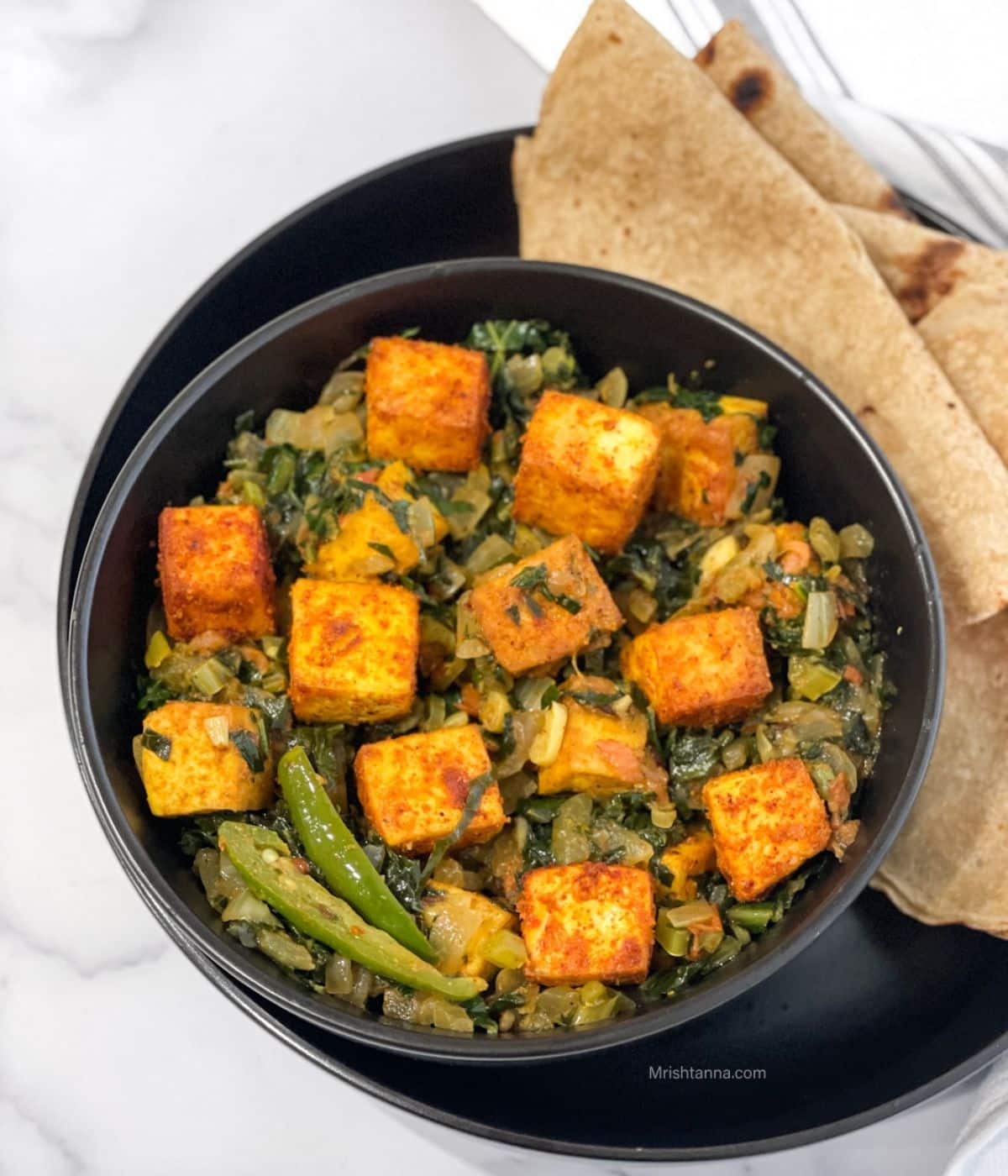 A plate is with chapati and bowl of vegan saag paneer on the table.