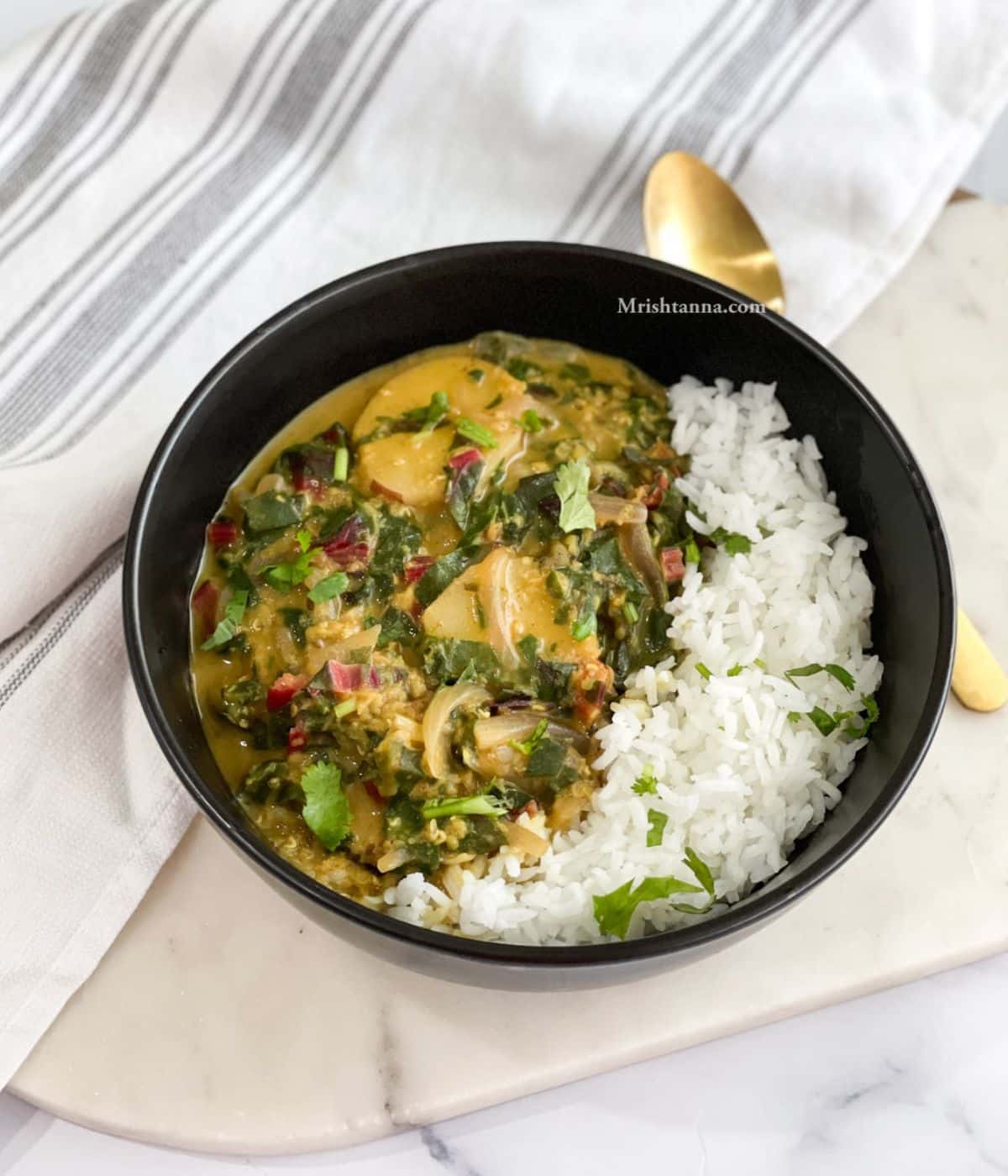 A bowl of swiss chard curry and rice is on the white marble tray and with a golden spoon and cloth napkin.