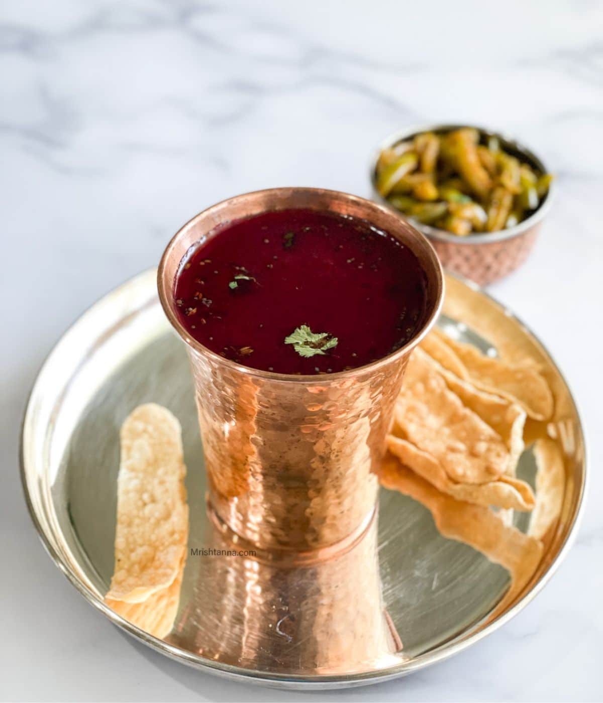 A copper mug is filled with beetroot rasam on the plate along with papad.