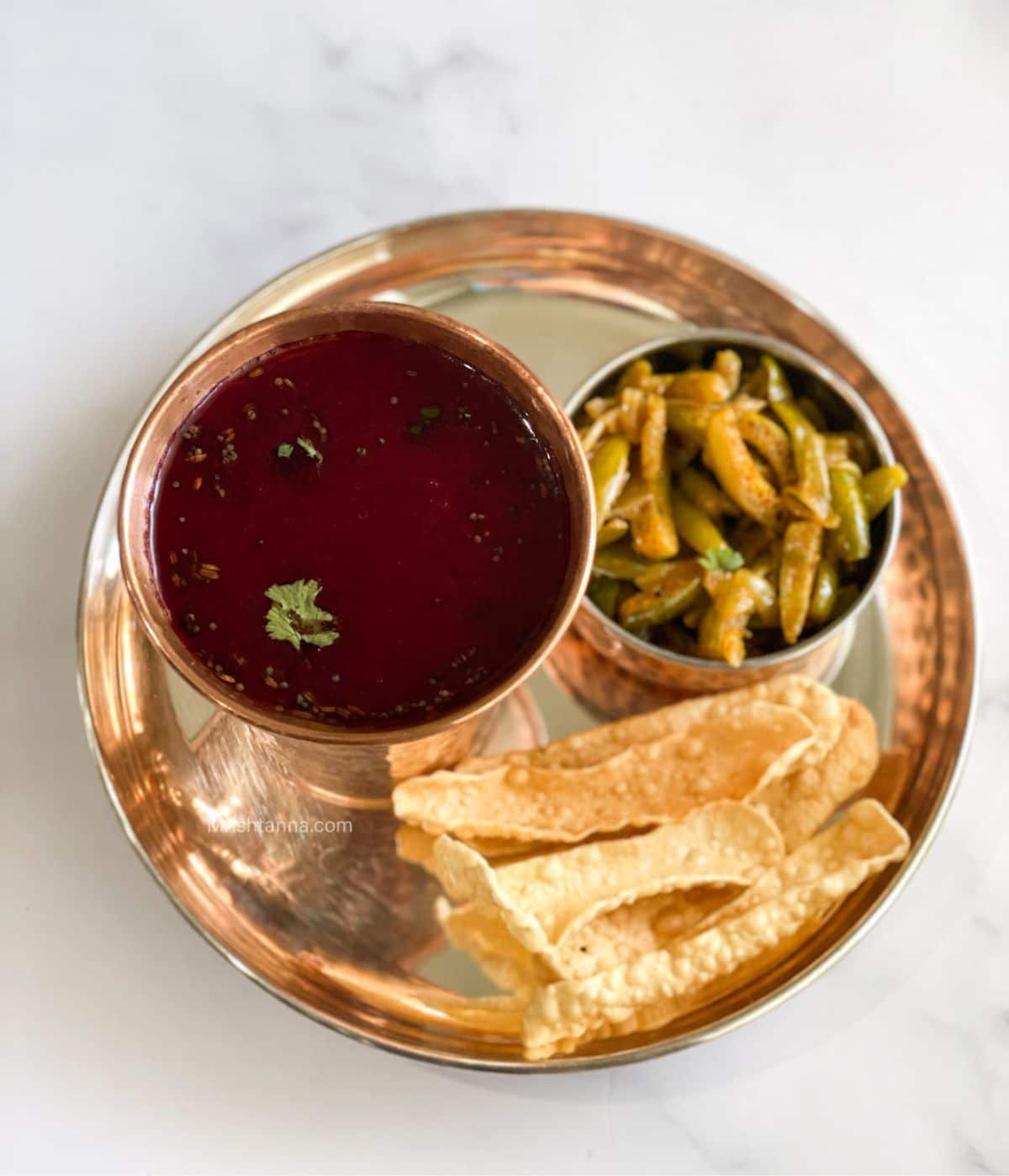 A copper glass is filled with beetroot rasam and placed on the silver plate with stir fry.