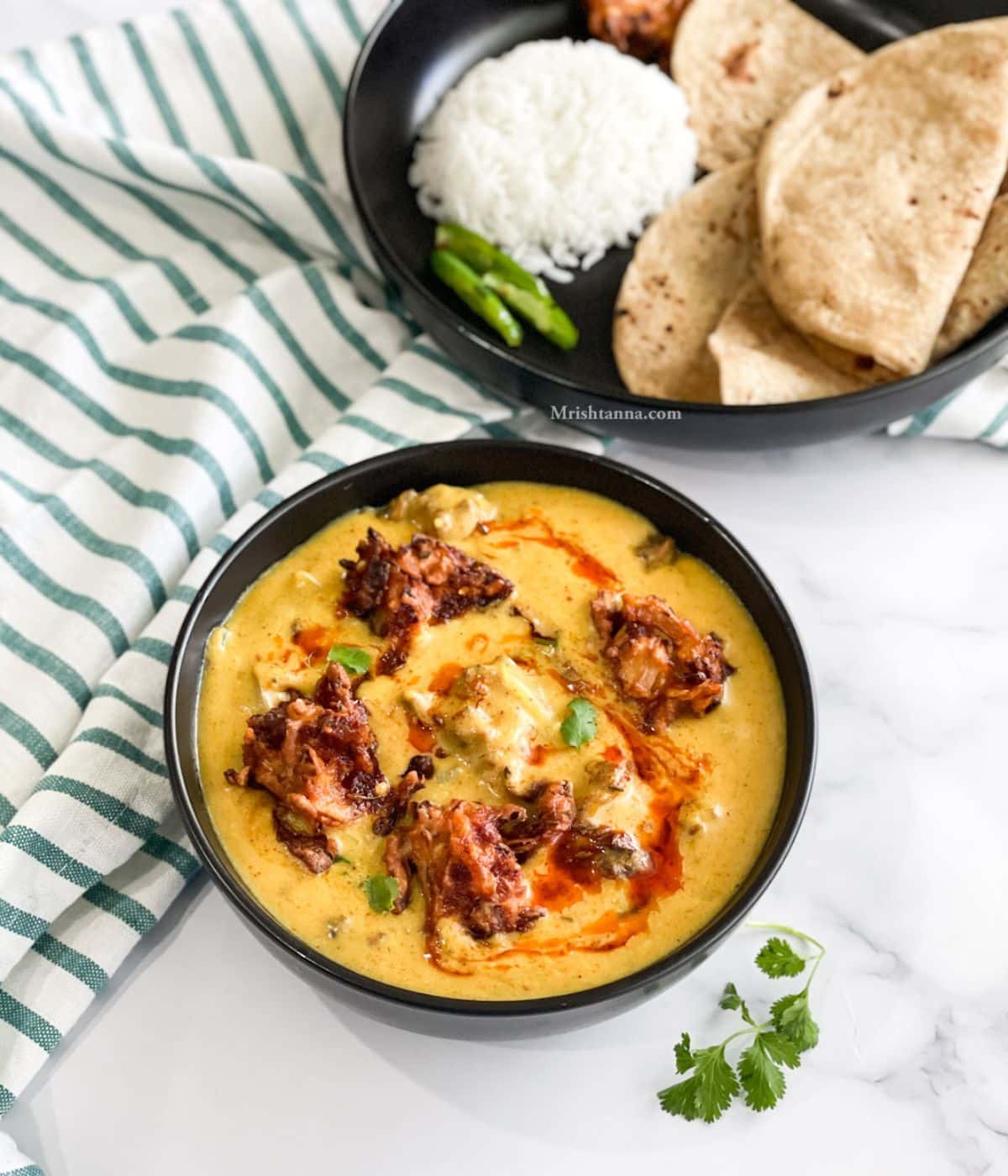 A bowl of kadhi pakora curry is on the table with a plate of rice and chapati.