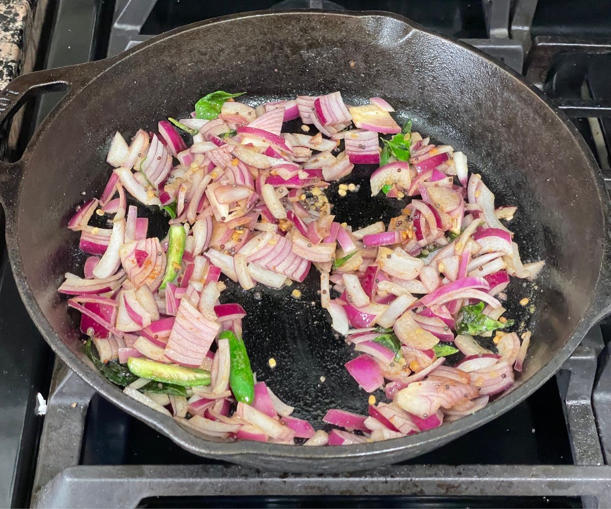 Cast iron pan is with spices, green chilies and chopped onions over the flame.
