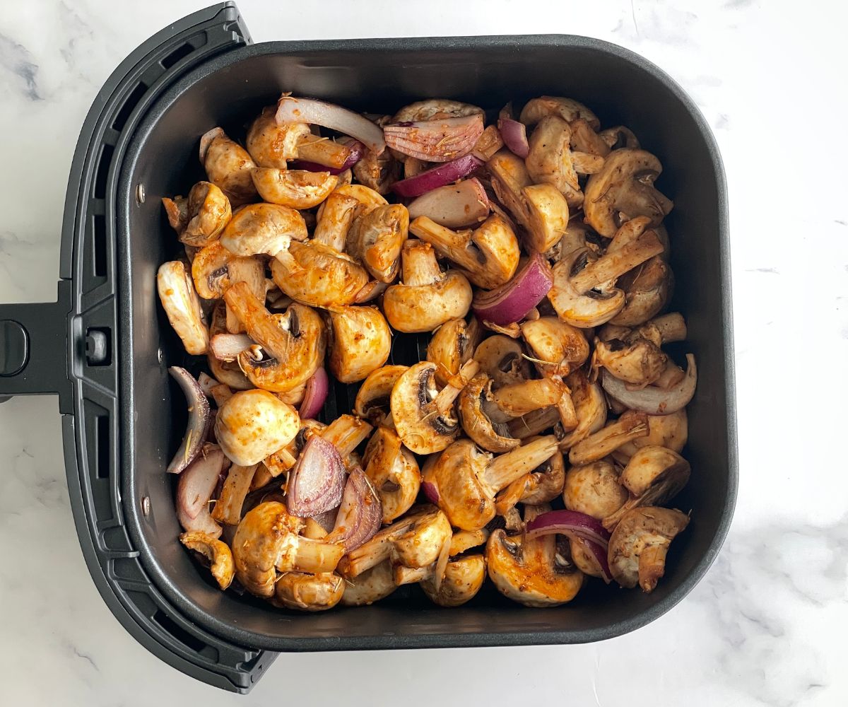 Air fryer basket is with coated mushrooms and onions.