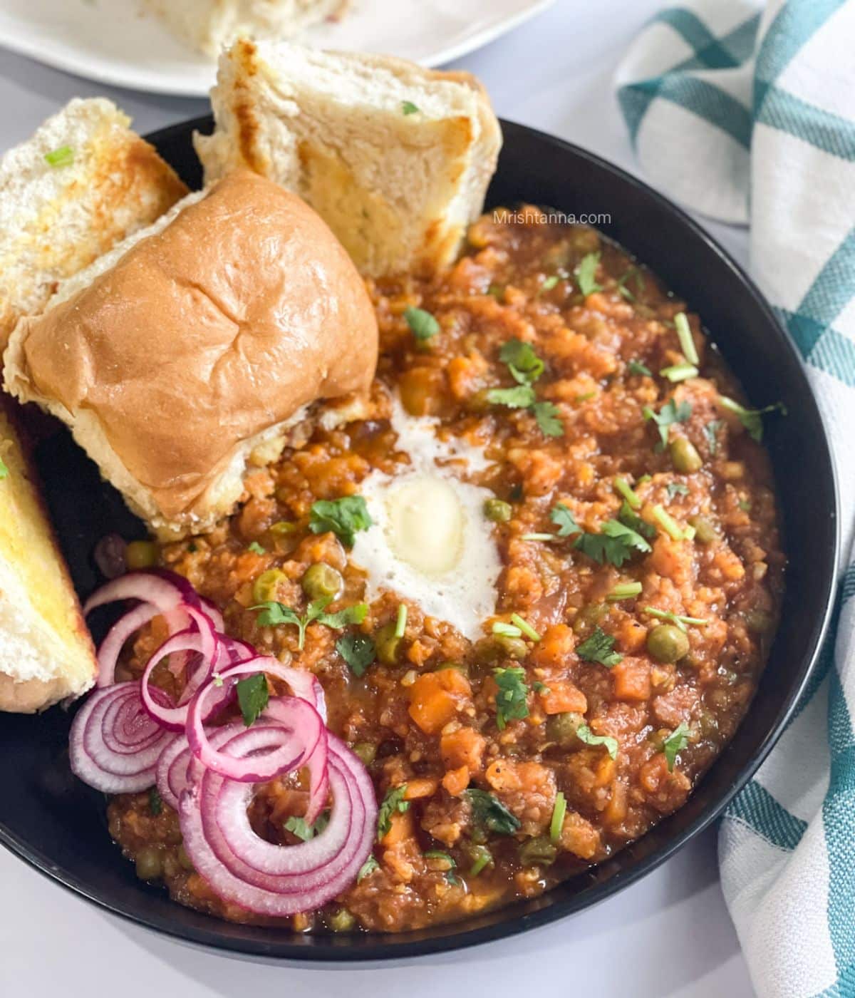 A plate of pav bhaji is on the table topped with vegan butter and sliced onions.