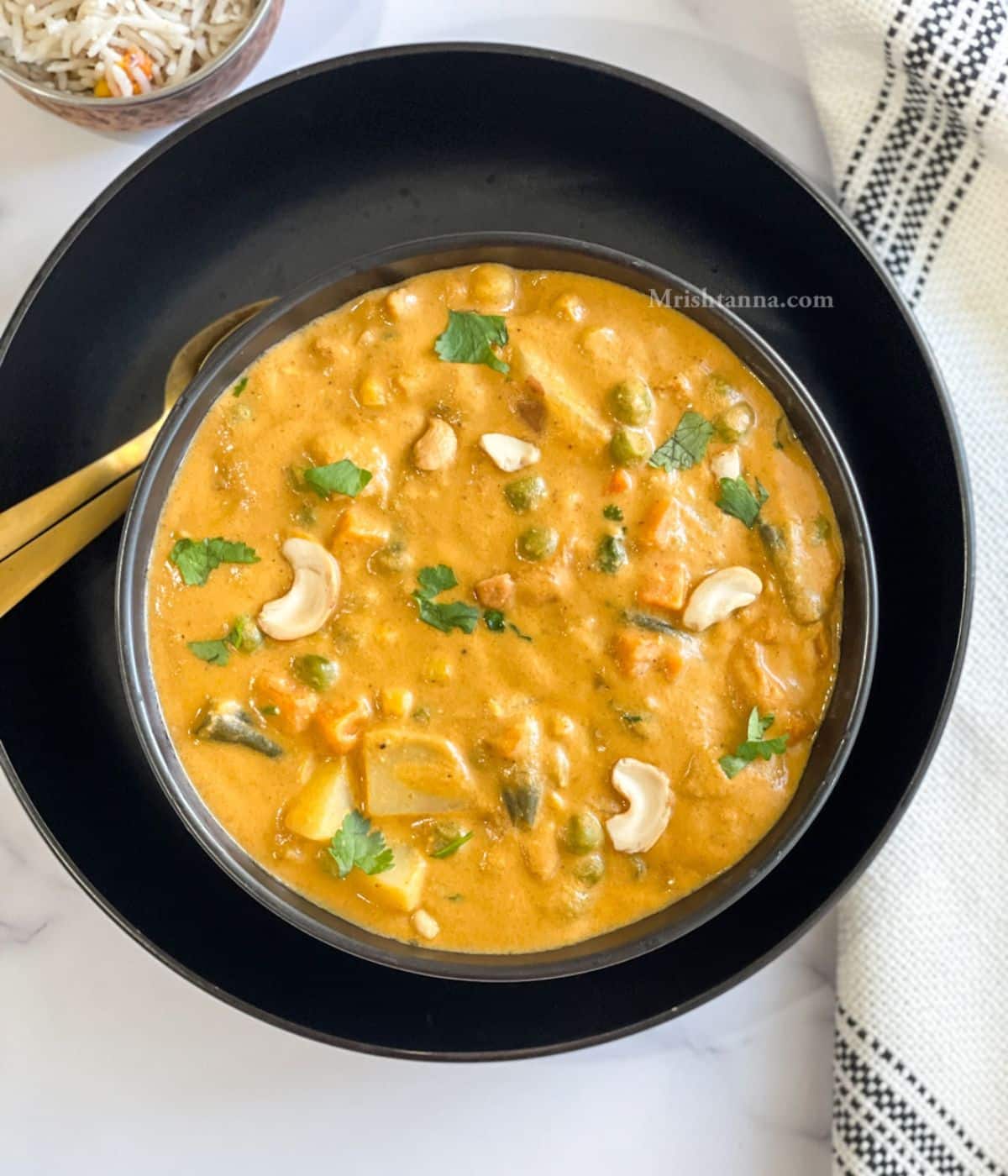 A bowl of instant pot vegetable korma is on the black plate with golden spoons.