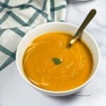 A bowl of vegan butternut squash carrot soup is on the table with a spoon inserted.