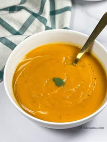 A bowl of vegan butternut squash carrot soup is on the table with a spoon inserted.