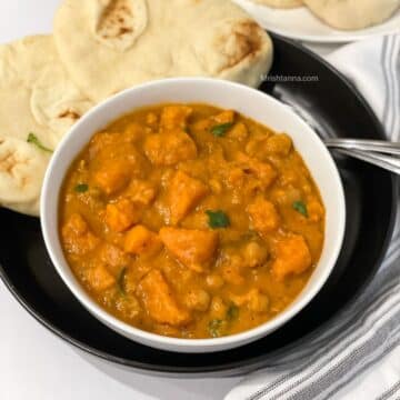 A bowl of sweet potato curry is on the black plate with spoons and naan.