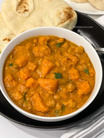 A bowl of sweet potato curry is on the black plate with spoons and naan.