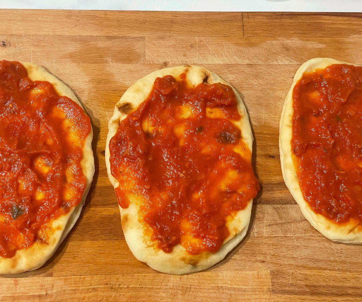 A wooden board is with naans and topped with pasta sauce.