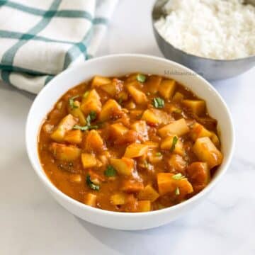 Bowl of potato tomato curry is placed on the white table.