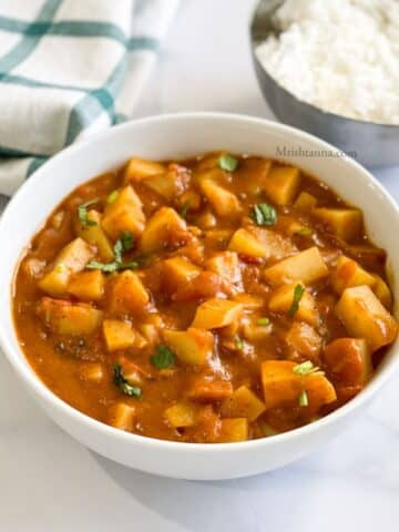 Bowl of potato tomato curry is placed on the white table.