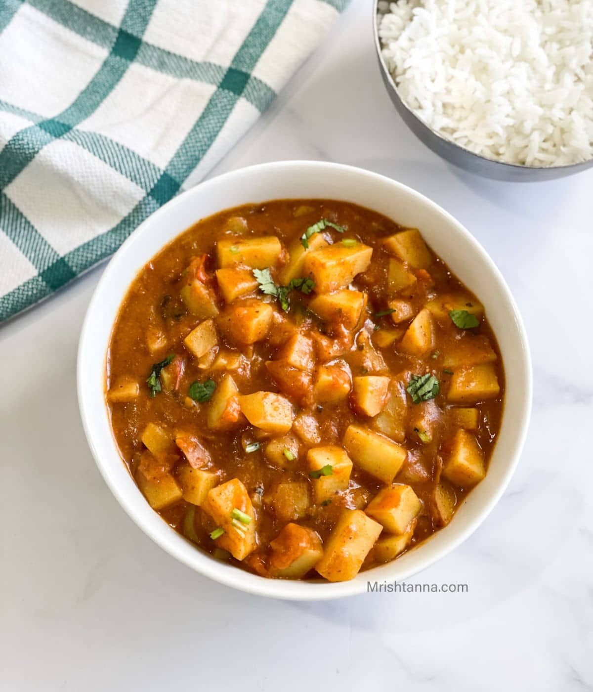A bowl of aloo tomato curry is on the table with a side of rice.