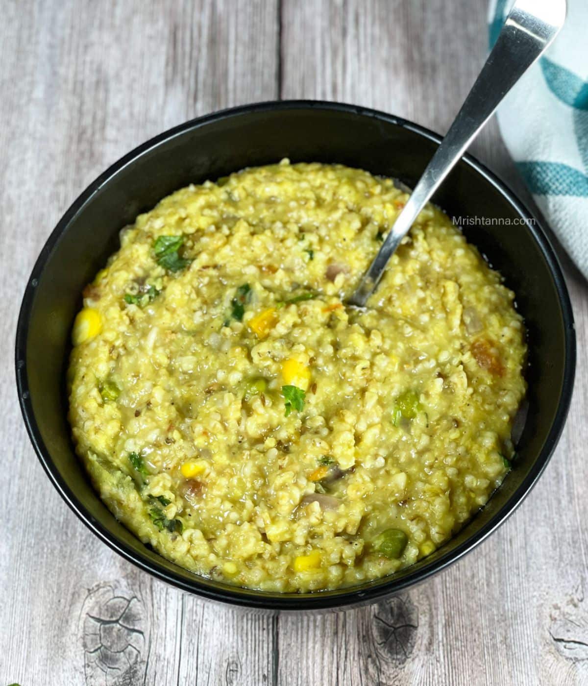 A bowl of oats khichdi is on the table with a spoon inserted.