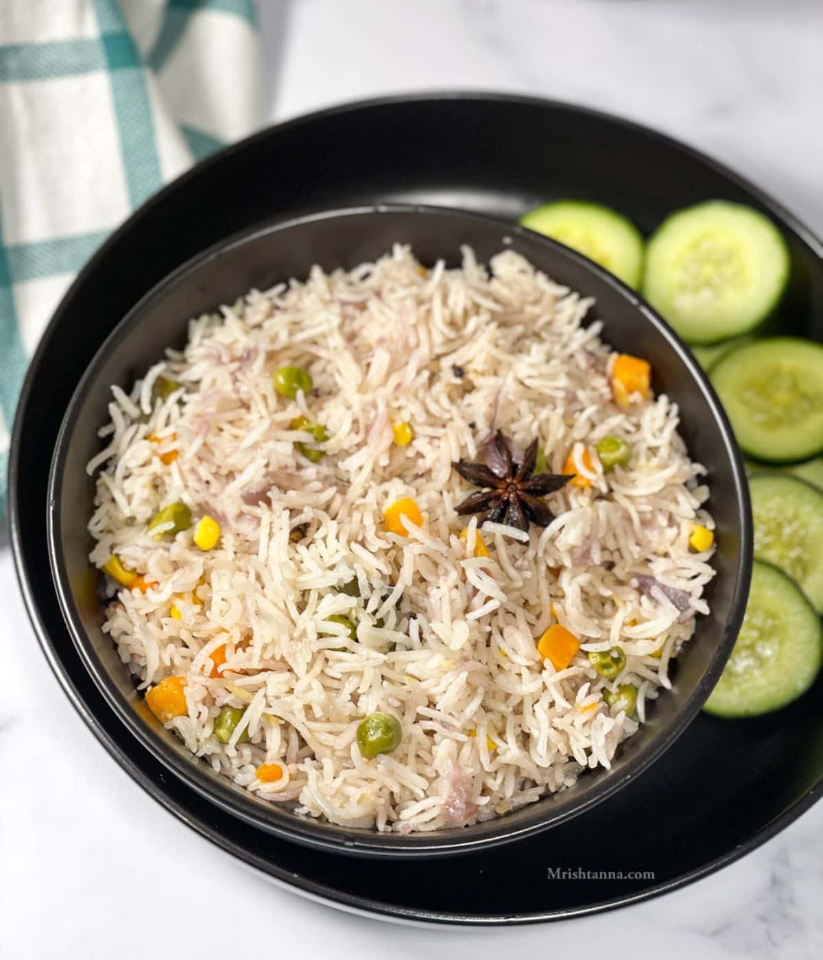 A bowl of vegetable pulao is on the plate along with cucumber slices.