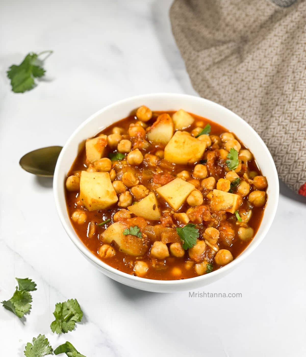 A bowl of aloo chana masala is on the table with a spoon by the side.