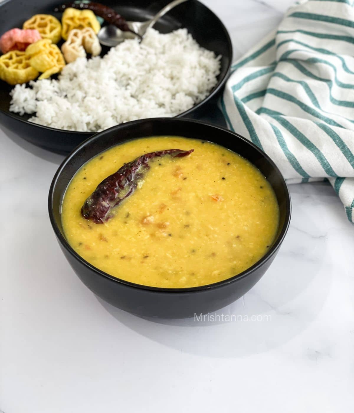 A bowl of bengali moong dal is on the table with a plate of rice.