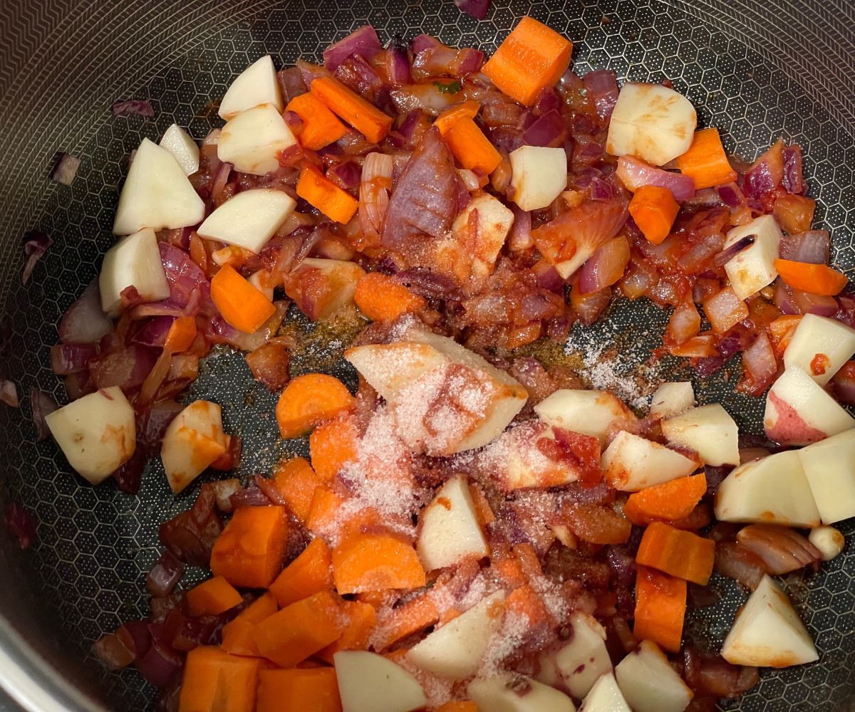 A pan is with onions, tomato paste, potatoes and carrots over the heat.