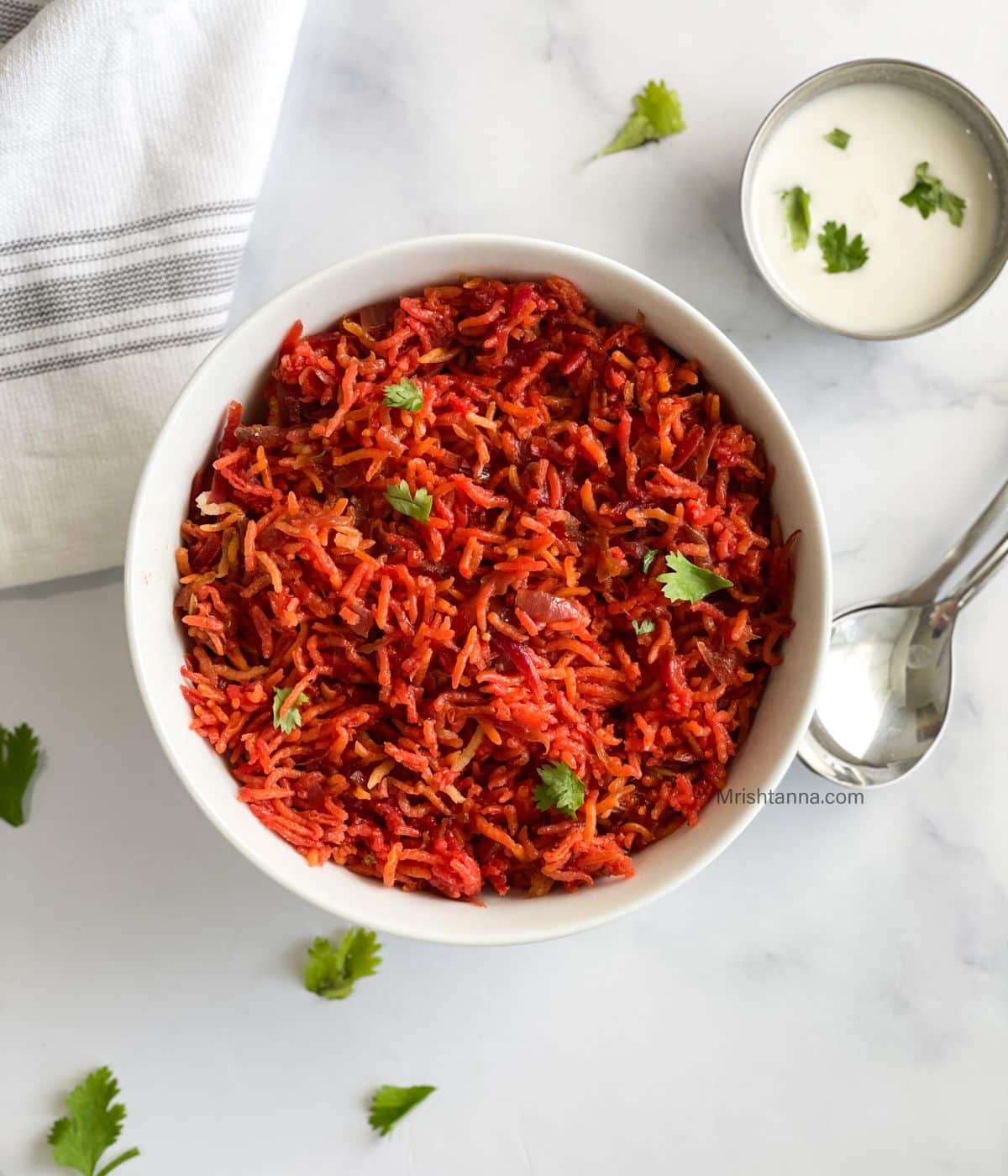 A bowl of beetroot rice is on the table along with a spoon and bowl of raita.