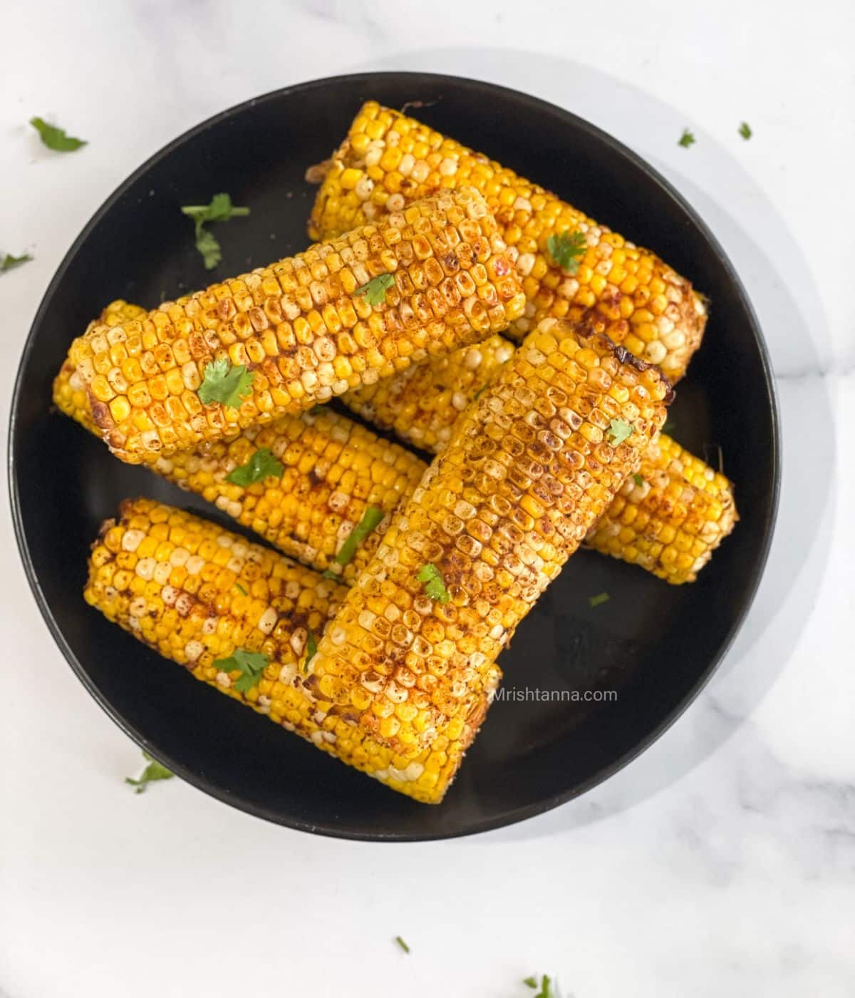 A plate of corn on the cob is on the table topped with spices and herbs.