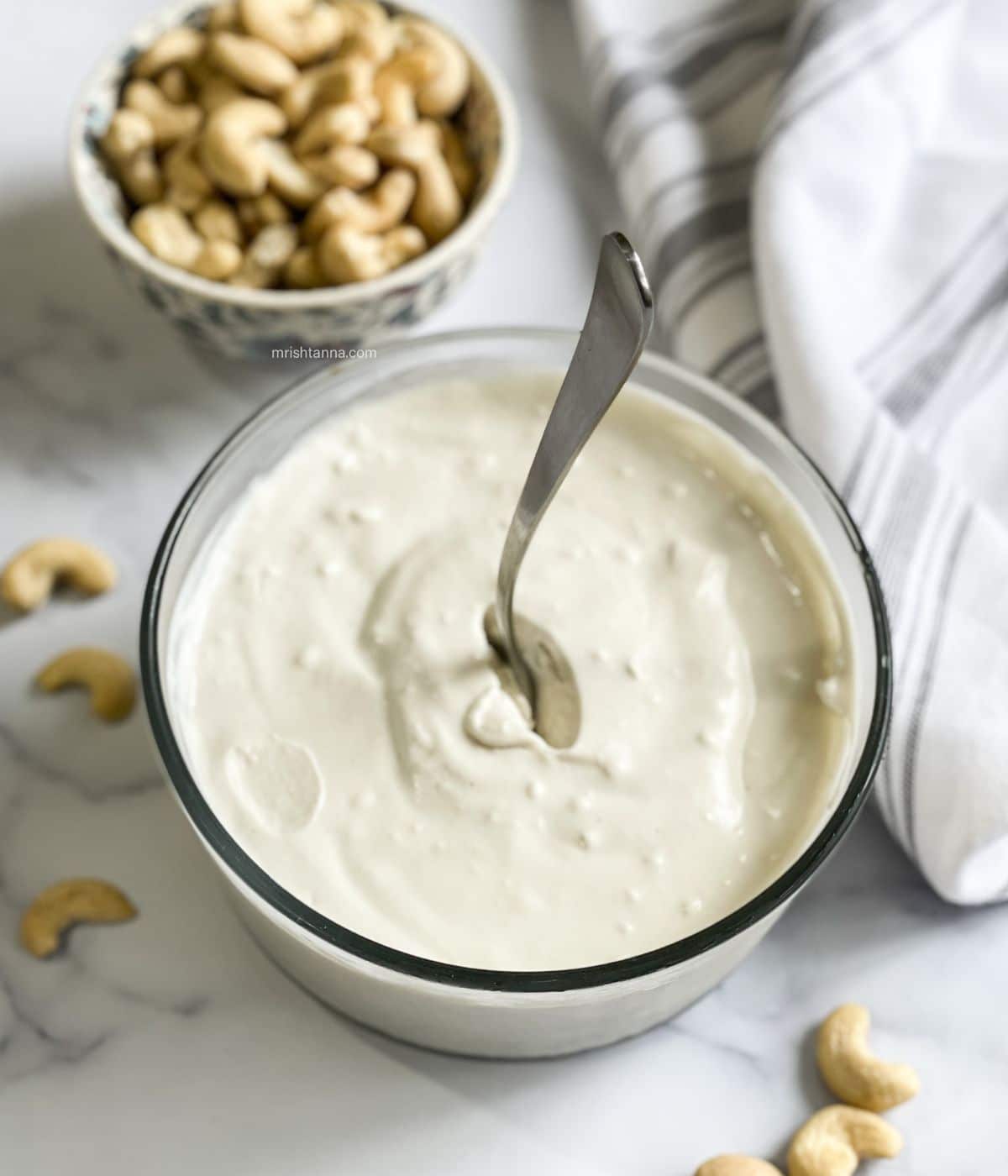 A glass bowl has cashew yogurt and in the center a spoon is inserted.