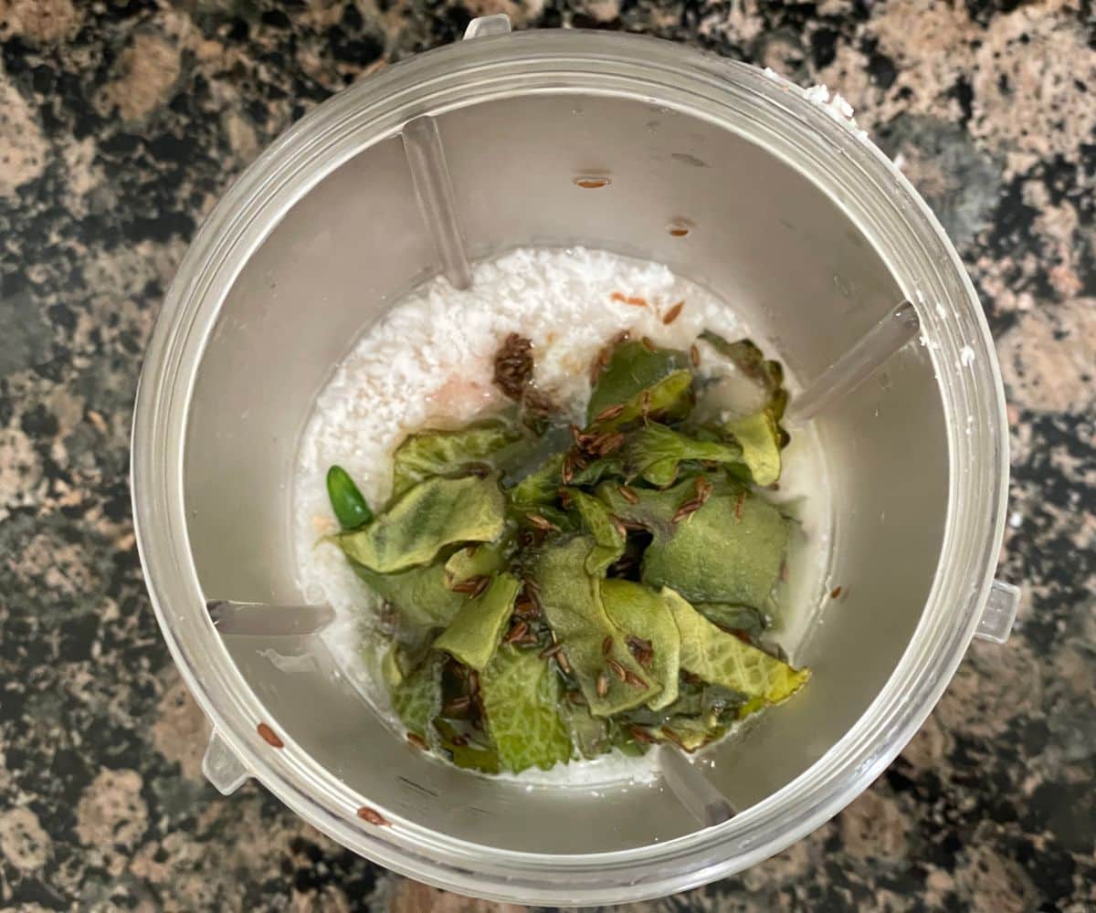 A blender is filled with coconuts, spices and sauteed Indian borage leaves.