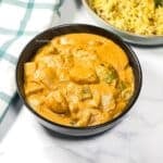 Bowl of creamy orange potato tikka masala on a white marble surface next to a green‑striped kitchen towel and a plate of rice.