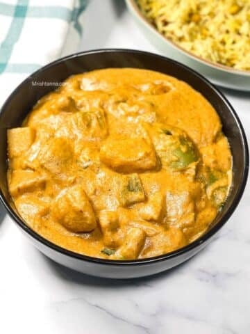 Bowl of creamy orange potato tikka masala on a white marble surface next to a green‑striped kitchen towel and a plate of rice.